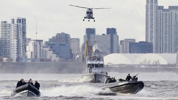 Ein Hubschrauber fliegt über zwei Schnellboote mit bewaffneten Soldaten vor einer Stadtkulisse.