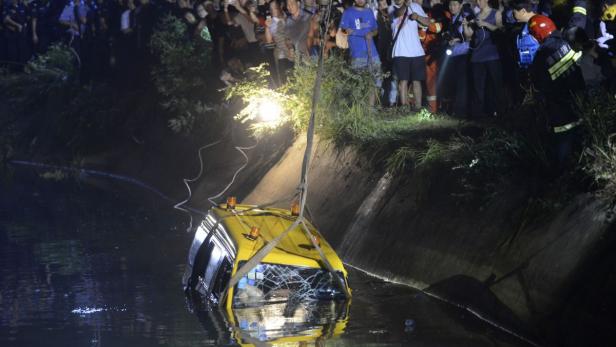 Ein gelbes Auto liegt in einem Kanal, während Menschen am Ufer zusehen.