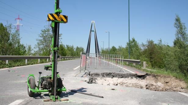 Eine Straßenbaustelle mit einem Bohrgerät und einer Brücke im Hintergrund.