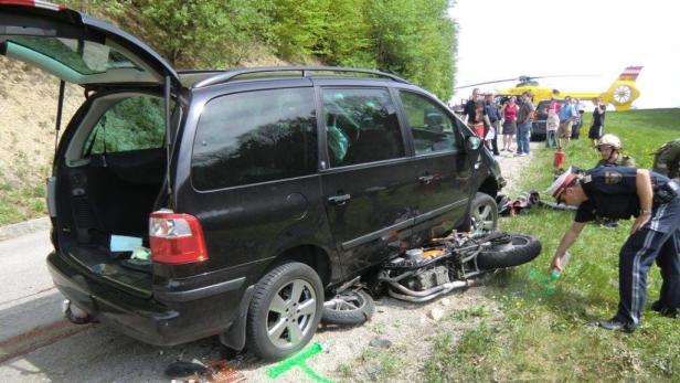 Ein Motorrad liegt unter einem schwarzen Auto, während ein Polizist den Unfallort untersucht. Im Hintergrund ein Rettungshubschrauber.