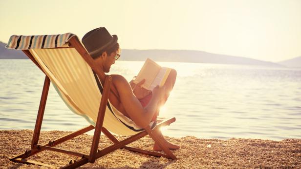 Ein Mann mit Hut liest in einem Liegestuhl am Strand ein Buch.