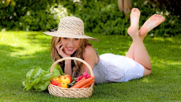 Eine Frau mit Hut liegt barfuß auf einer Wiese neben einem Korb mit Gemüse und Obst.