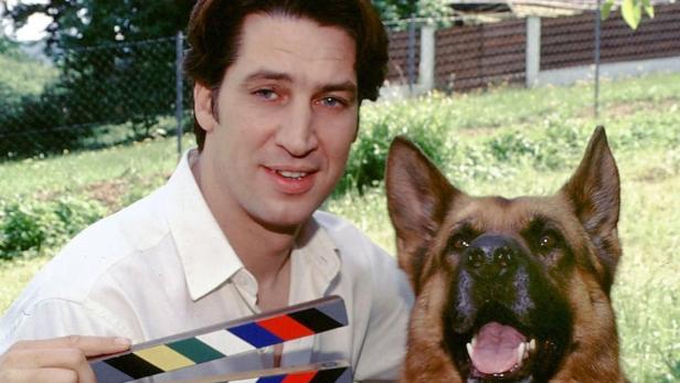 Ein Mann mit Filmklappe posiert mit einem Schäferhund im Freien.