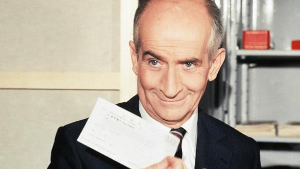 Ein Mann im Anzug hält einen Scheck in der Hand und lächelt verschmitzt.