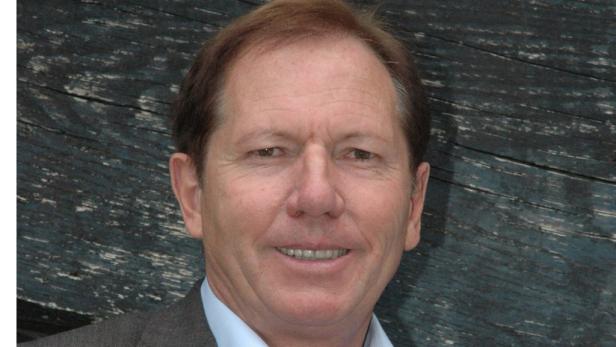 Ein Mann mit braunen Haaren lächelt vor einem Hintergrund aus dunklem Holz.