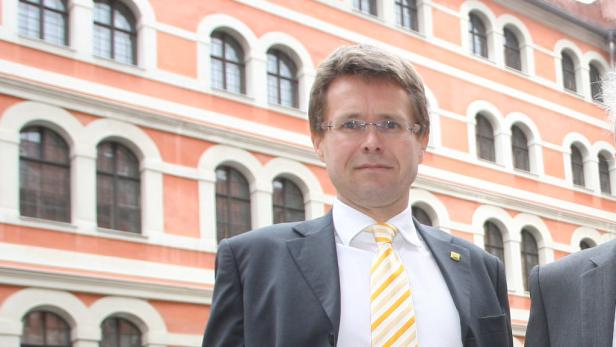 Ein Mann mit Brille und Anzug steht vor einem Gebäude mit vielen Fenstern.