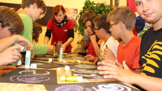 Eine Gruppe Kinder bastelt mit Glitzer und Klebstoff an Tischen.