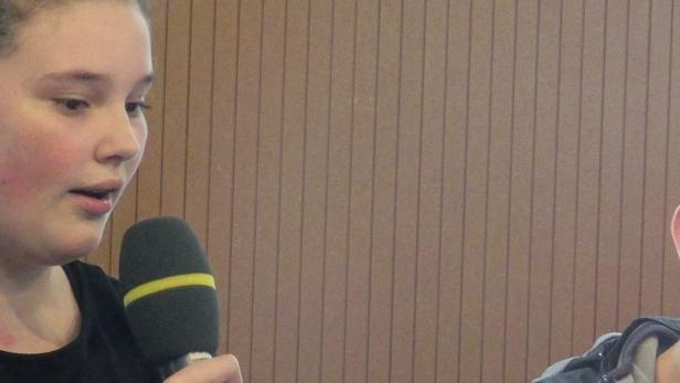 Ein Mädchen spricht in ein Mikrofon, während ein Junge im Hintergrund lächelt.