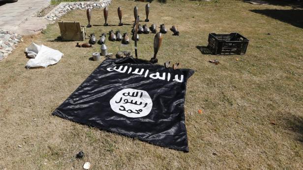 Eine Flagge des Islamischen Staates liegt auf einer Wiese, umgeben von Mörsergranaten.