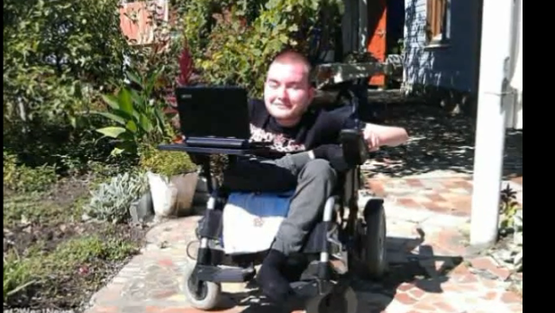 Ein junger Mann sitzt im Rollstuhl im Garten und benutzt einen Laptop.