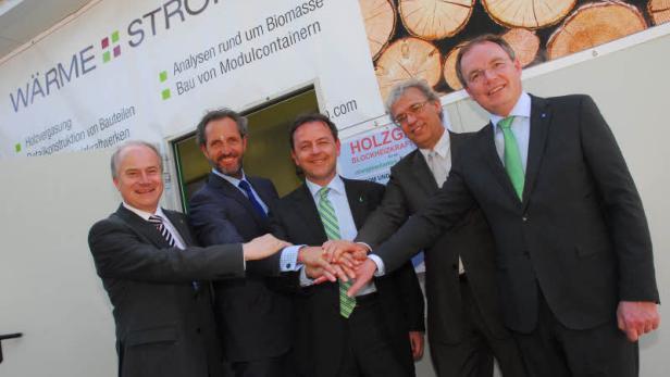 Fünf Männer in Anzügen stehen vor einem Container mit der Aufschrift „Wärme Strom“ und stapeln ihre Hände.