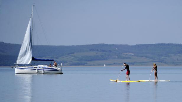 Zwei Personen paddeln auf dem See, im Hintergrund ein Segelboot.