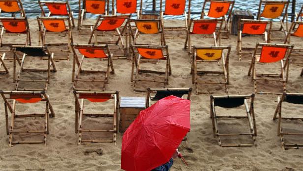 Ein roter Regenschirm steht zwischen vielen leeren Liegestühlen am Strand.