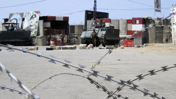 Militärfahrzeuge und Soldaten an einem Kontrollpunkt mit Stacheldraht im Vordergrund.