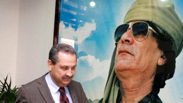 Ein Mann im Anzug steht neben einem Porträt von Muammar al-Gaddafi.