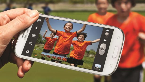 Ein Smartphone zeigt ein Foto von jubelnden Kindern in orangefarbenen Trikots auf einem Fußballfeld.