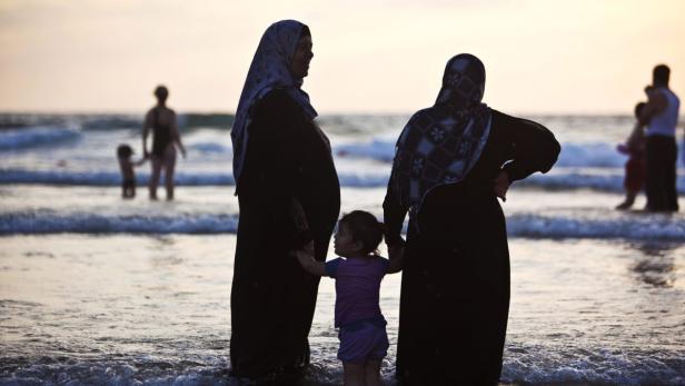 Zwei Frauen in traditioneller Kleidung stehen mit einem kleinen Mädchen am Strand im Wasser.