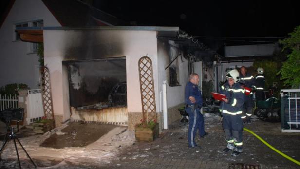 Nach einem Brand stehen Feuerwehrleute und ein Polizist vor einer beschädigten Garage.