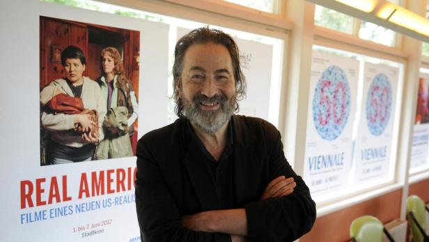 Ein Mann mit Bart steht vor einem Poster für die Filmreihe „Real American“.