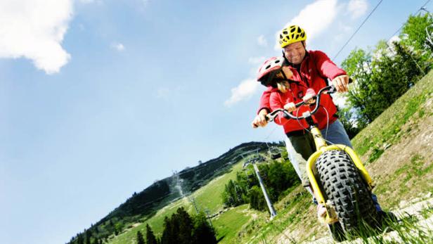 Ein Vater fährt mit seinem Kind auf einem Berg mit einem gelben Roller bergab.