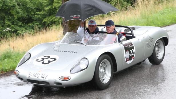 Zwei Männer fahren in einem silbernen Oldtimer-Rennwagen im Regen.