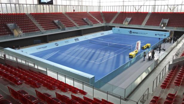 Ein leerer Tennisplatz mit blauer Oberfläche und roten Zuschauersitzen in einer Halle.