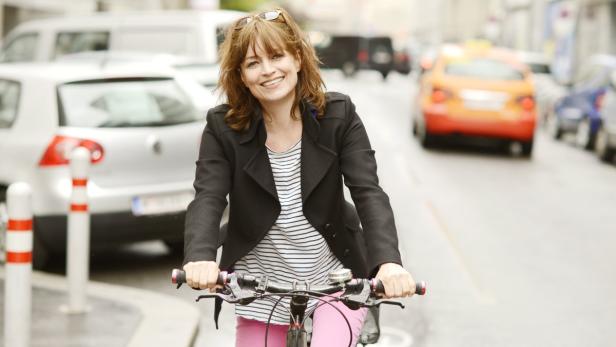 Eine Frau mit Jacke und rosa Hose fährt Fahrrad in der Stadt.