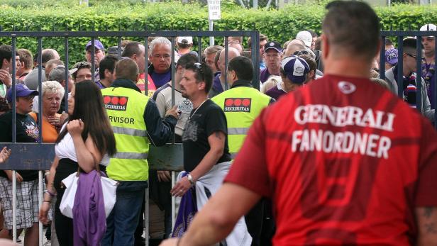 Eine Menschenmenge steht hinter einem Gitter, bewacht von Sicherheitskräften.