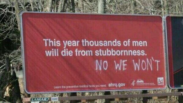 Eine rote Werbetafel warnt: „Dieses Jahr werden Tausende Männer an Sturheit sterben.“ Darunter steht gekritzelt: „NEIN, WERDEN WIR NICHT“.