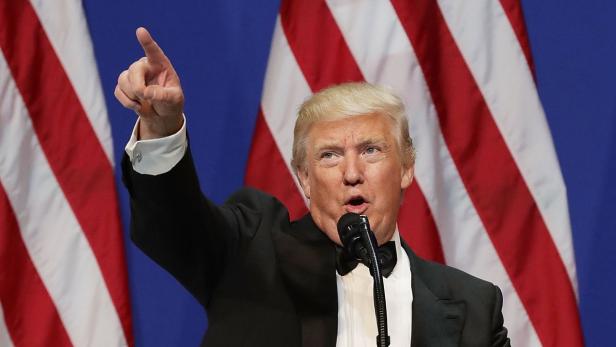 Donald Trump steht vor einer Fahne und hebt die Hand
