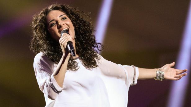 Eine Sängerin mit lockigem Haar singt in ein Mikrofon auf einer Bühne.