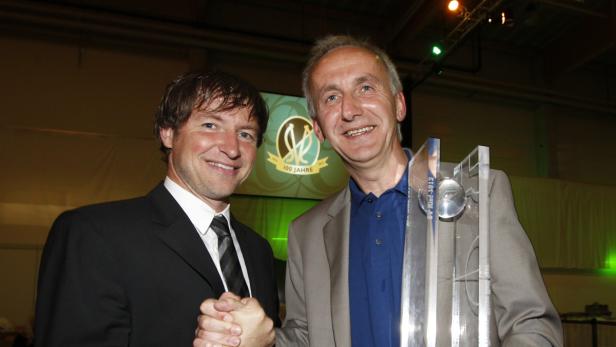 Zwei Männer posieren mit einer Trophäe vor einem Bildschirm mit dem Logo „100 Jahre SV Ried-3912“.