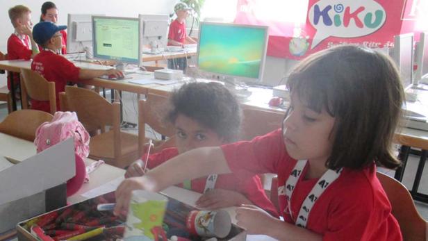 Kinder sitzen an Tischen und arbeiten an Computern und mit Stiften in einem Klassenzimmer.