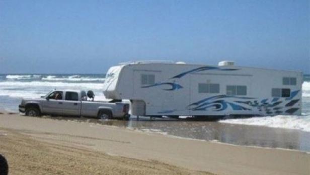 Ein Wohnwagen wird von einem Pickup-Truck am Strand ins Meer gezogen.