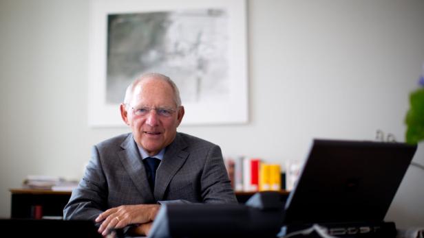 Ein älterer Mann mit Brille und Anzug sitzt an einem Schreibtisch mit Laptop.