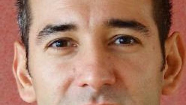 Ein Porträt eines Mannes mit dunklen Haaren und braunen Augen vor einem roten Hintergrund.