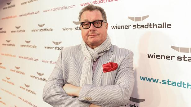 Ein Mann mit Brille, Schal und gestreiftem Sakko posiert vor einem Wiener Stadthalle-Hintergrund.