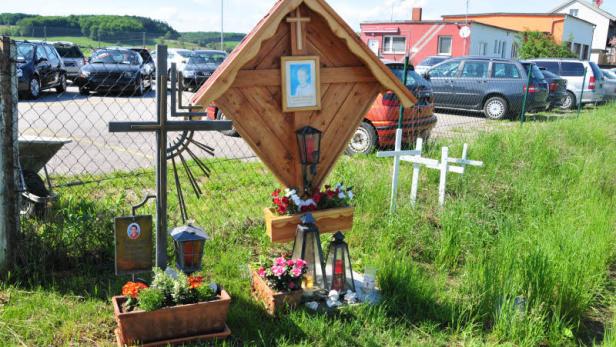 Ein hölzernes Wegkreuz mit Blumen und Gedenklichtern am Straßenrand.