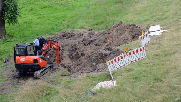 Ein Bagger hebt Erde aus, während zwei Männer daneben stehen und die Arbeiten beobachten.