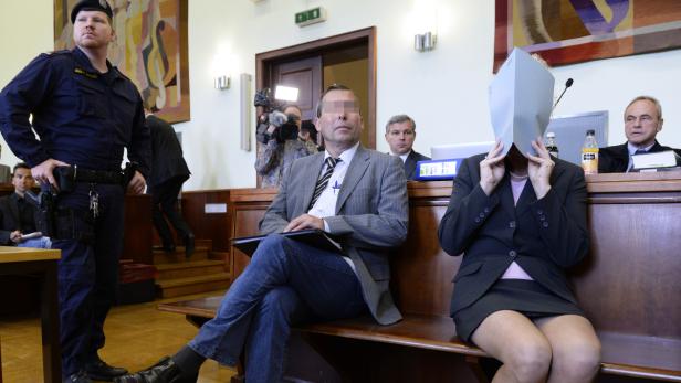 In einem Gerichtssaal sitzen ein Mann und eine Frau, die ihr Gesicht mit einem Blatt Papier bedeckt.