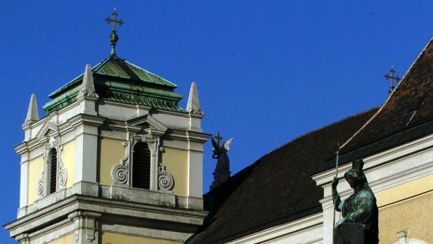 Detailansicht eines gelben Kirchturms mit Statuen und einem Kreuz gegen einen blauen Himmel.