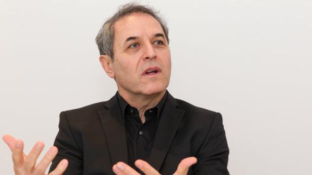 Ein Mann in einem schwarzen Anzug gestikuliert vor einem weißen Hintergrund.