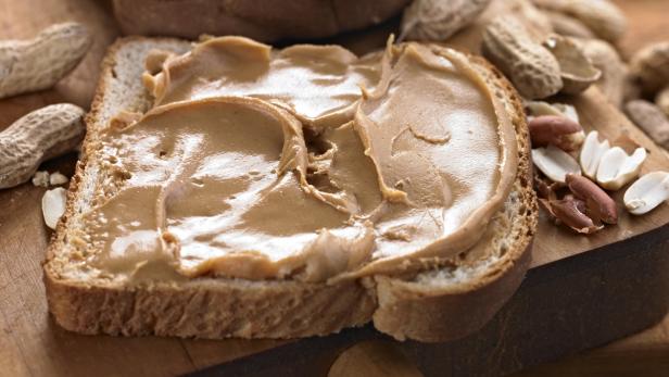 Ein Erdnussbutterbrot liegt auf einem Holzbrett, umgeben von Erdnüssen.