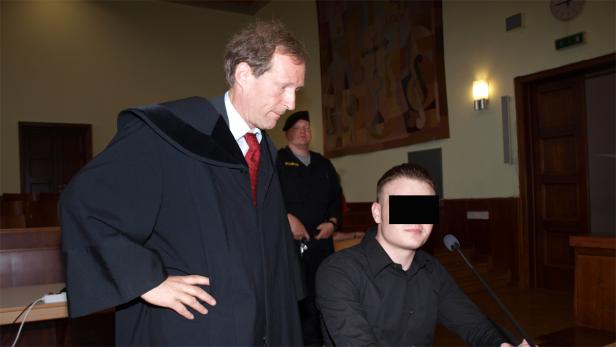 Ein Richter in Robe steht neben einem jungen Mann mit verdecktem Gesicht in einem Gerichtssaal.