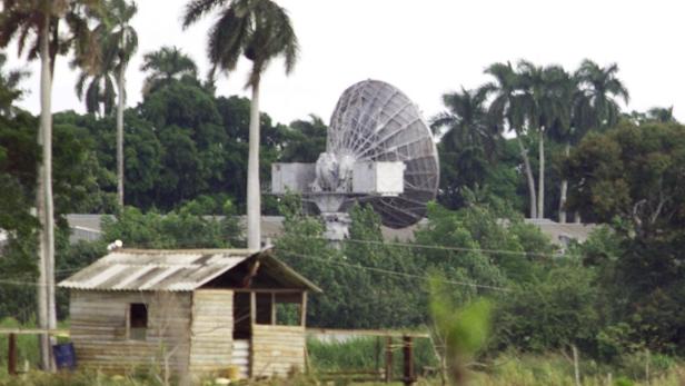 Eine große Satellitenschüssel hinter einem kleinen Holzhaus und Palmen.