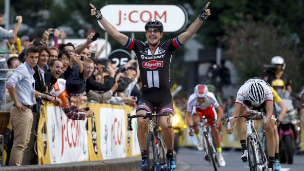 Ein Radrennfahrer überquert mit erhobenen Armen die Ziellinie, während die Zuschauer jubeln.