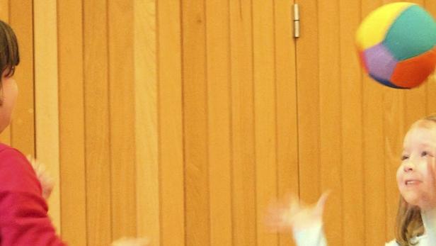 Zwei Kinder spielen mit einem bunten Ball vor einer Holzwand.