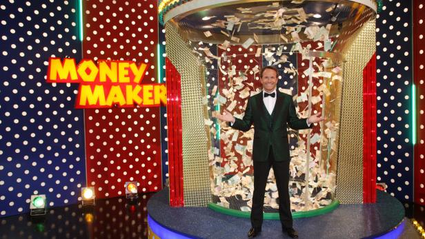 Ein Mann in einem grünen Anzug steht vor einer Maschine, in der Geldscheine herumwirbeln.