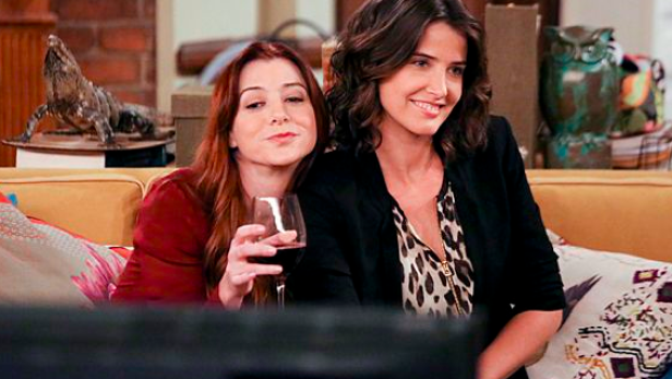 Alyson Hannigan und Cobie Smulders sitzen auf einer Couch und schauen fern.
