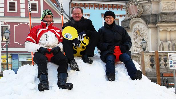 Drei Personen sitzen mit Rodeltellern auf einem Schneehügel.